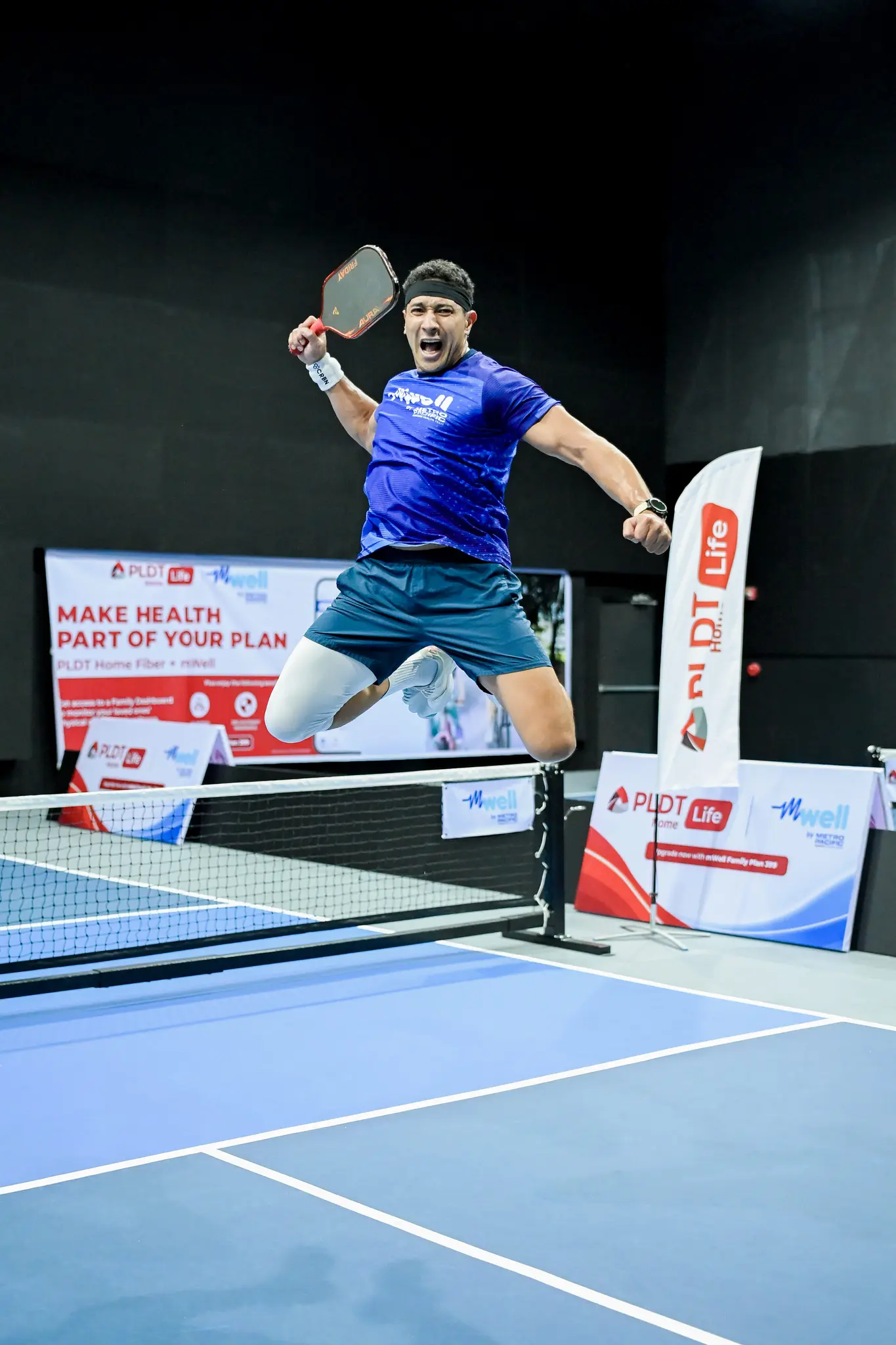A pickleball player in a blue mWell shirt jumps with a racket at a PLDT Home Life and mWell wellness event.